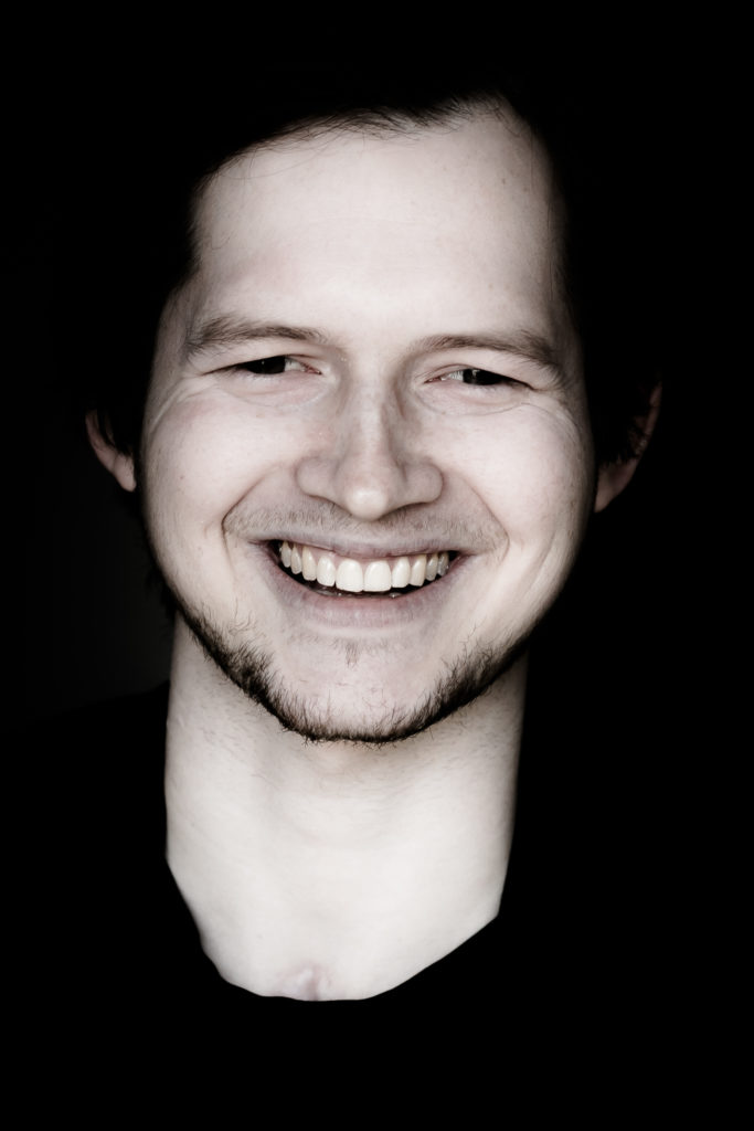 Porträtfoto von Simon Nickel - gleiche Bildbeschreibung wie weiter oben im Text: Junger Mann mit freundlichem, offenem Lächeln, dunklen Haaren, leichtem Bartansatz und schwarzem T-Shirt.