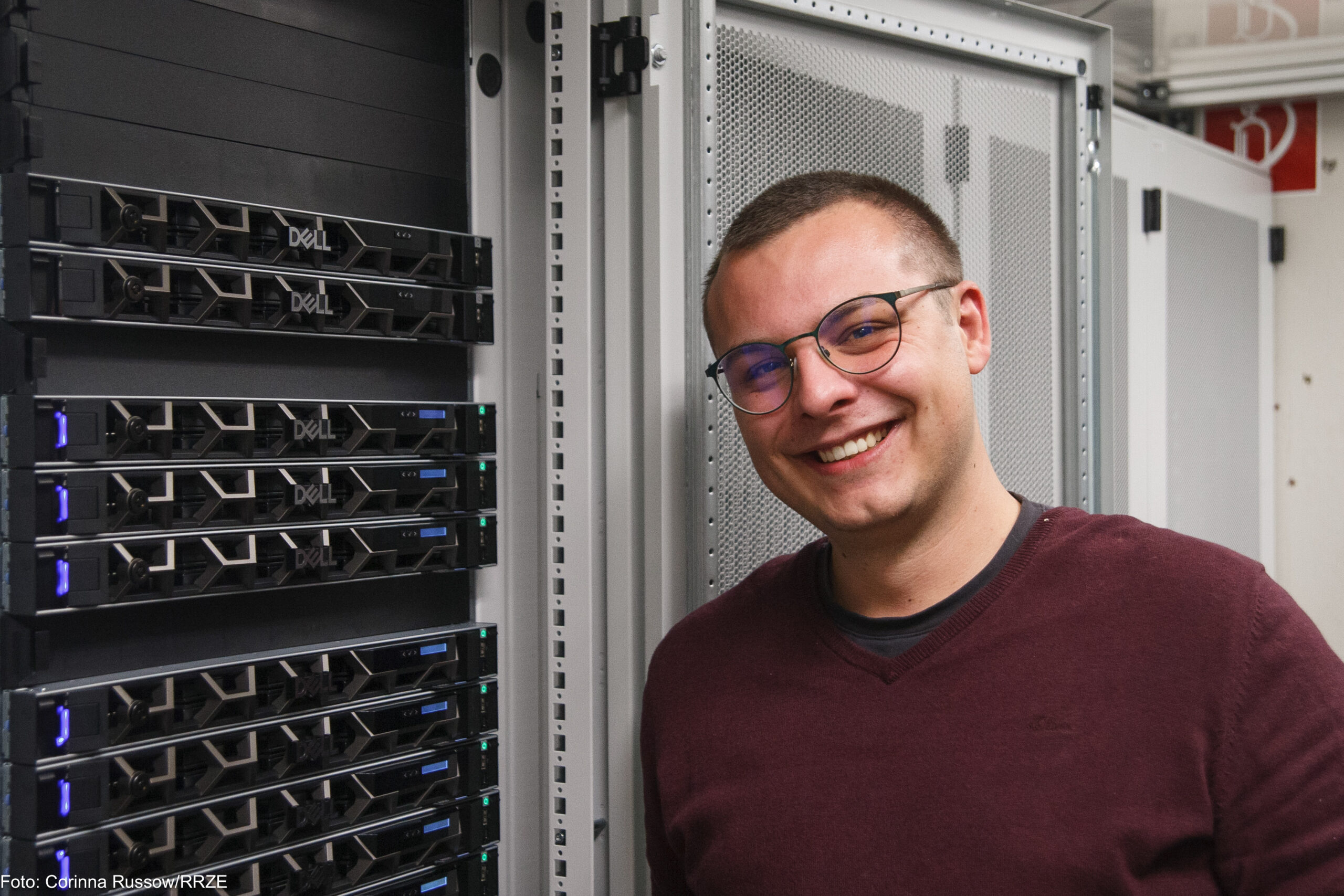 Ein junger Mann steht lächelnd in einem dunkelroten Pulli vor einem Schrank mit Servern. Die Server haben eine Gittertür, die offen steht, damit die schwarzen Server zu sehen sind.